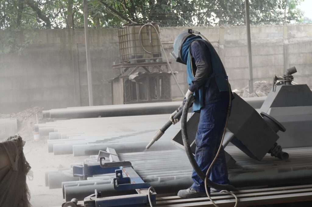 Pekerja sandblasting lengkap dengan APD melakukan proses sandblasting pada permukaan pipa industri untuk menghilangkan karat dan kotoran